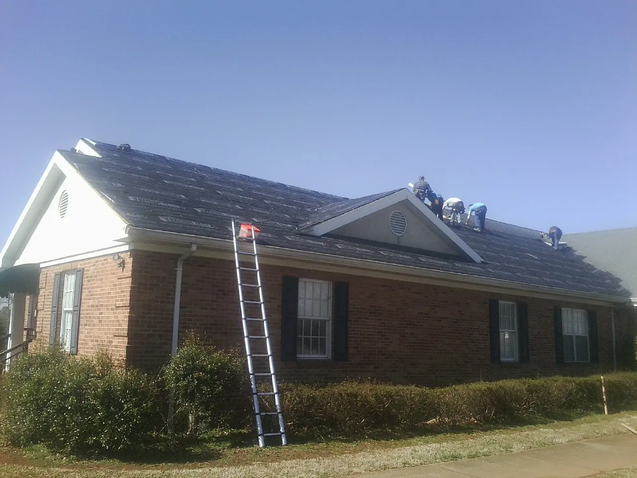 Crew tearing shingles off a brick ranch roof with a ladder set — Metal Roof Repair work by our crew in Lonsdale
