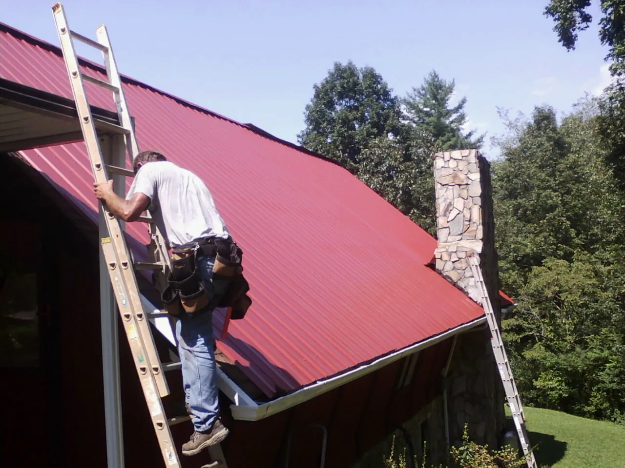 Roofer on a ladder beside a red metal roof and stone chimney — Soffit & Fascia Repair by Roofing Brothers in Lonsdale
