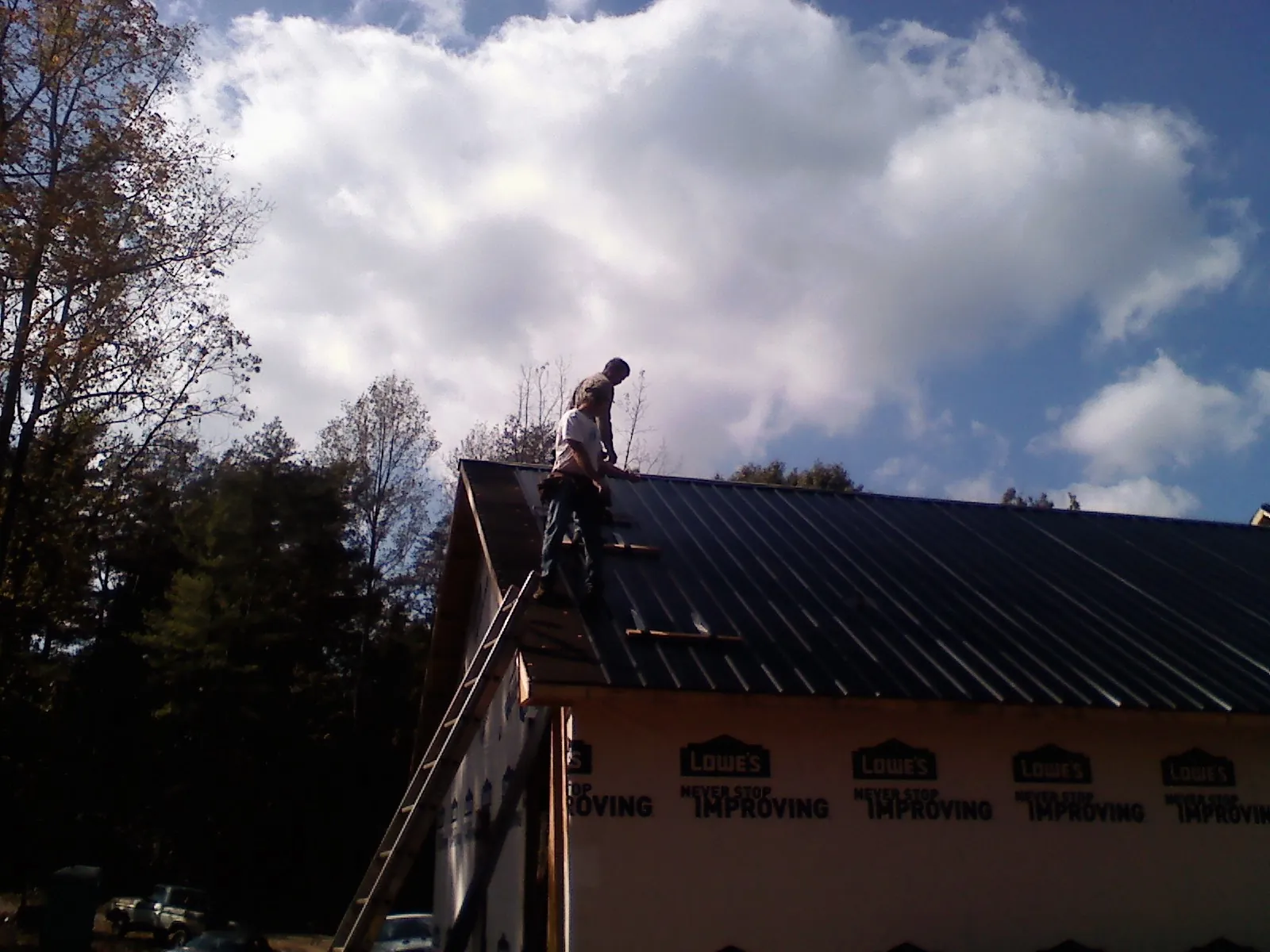 Roofer fastening dark metal panels high against a cloudy sky, from a Roof Inspection job in Lonsdale